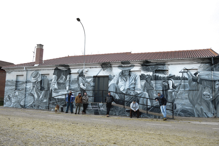 Jose, Víctor, Alfonso, Juanmi y los dos Rubén en la fachada de su sede, las viejas escuelas, con un precioso mural de faenas del campo, obra de dados.cero. | F.F.