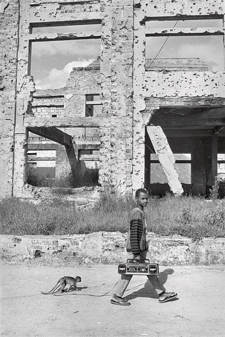 Un niño pasea a su mono entre las ruinas de su ciudad_Kuito (Angola), abril 1997. | GERVASIO SÁNCHEZ