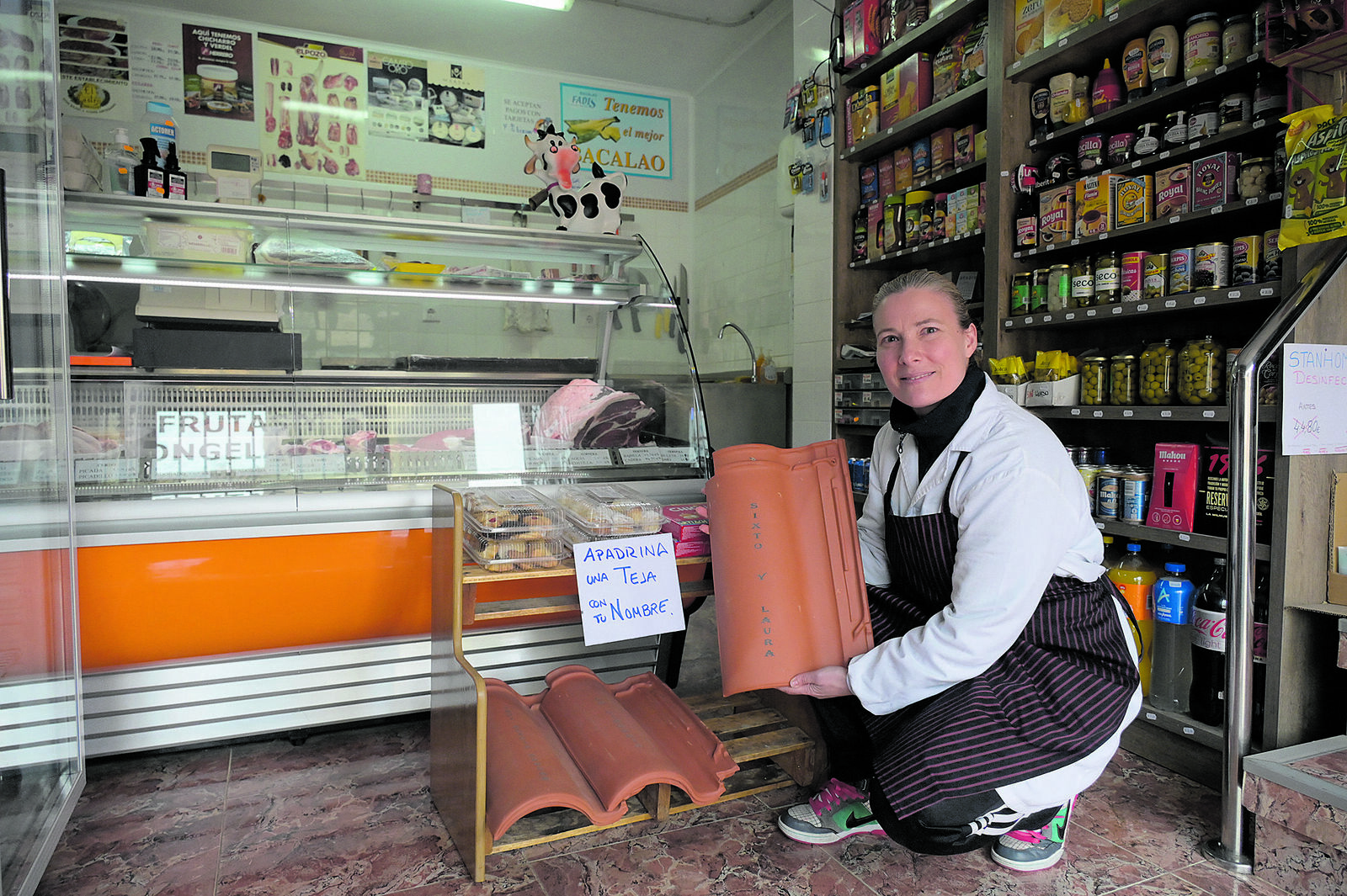 Laura Alegre, en el interior de su carnicería, posa con las tejas que tiene expuestas para animar a la gente a sumarse a la campaña. | MAURICIO PEÑA
