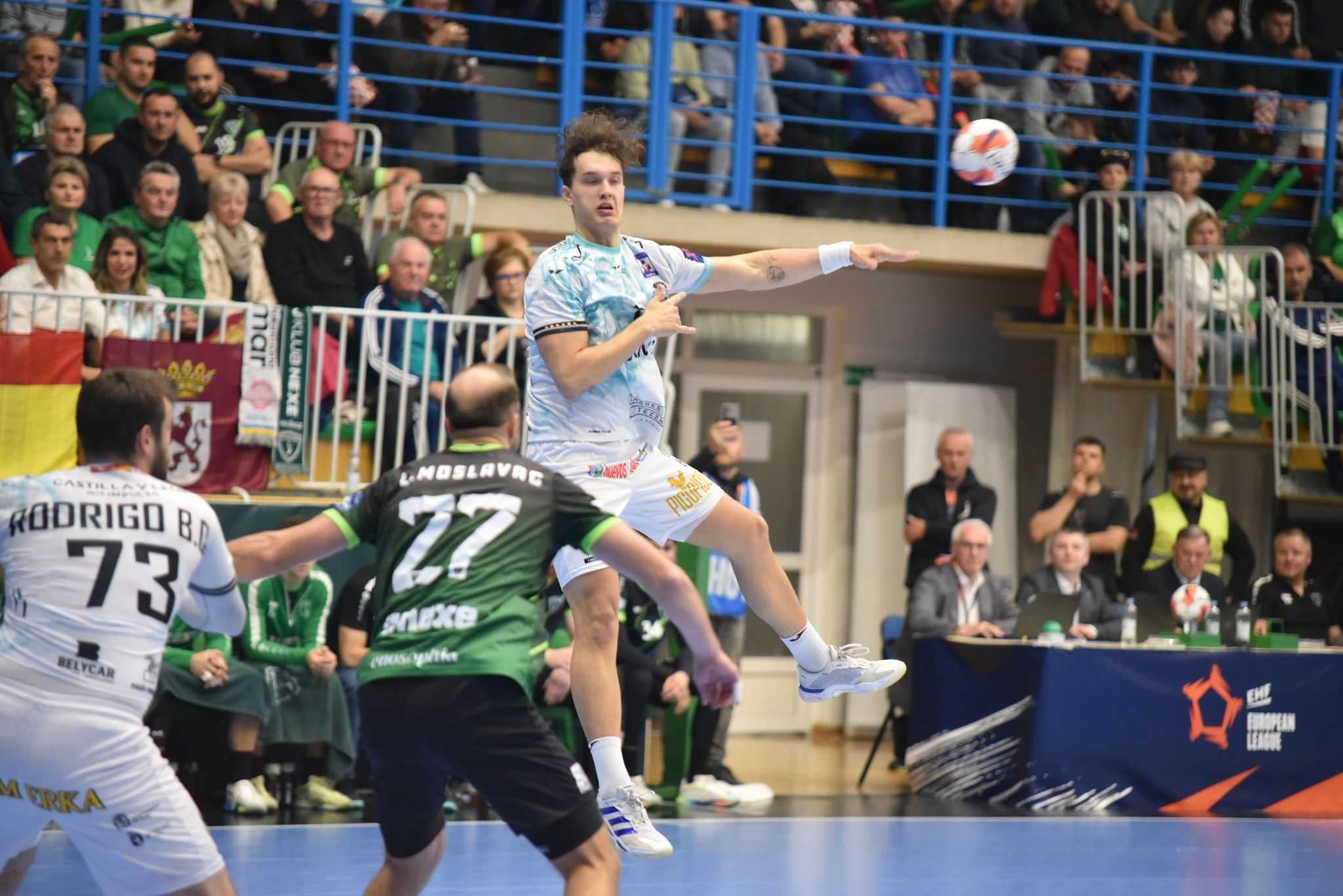 Wasiak da un pase en el aire conLuka Moslavac defendiendo y Benites pidiéndola en el pivote, mientras en la grada se ve la bandera de León. RK NEXE