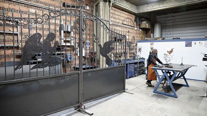 El artista y artesano a la derecha, ante su mesa de trabajo, y el portón del cementerio de Garfín a la izquierda El artista y artesano a la derecha, ante su mesa de trabajo, y el portón del cementerio de Garfín a la izquierda