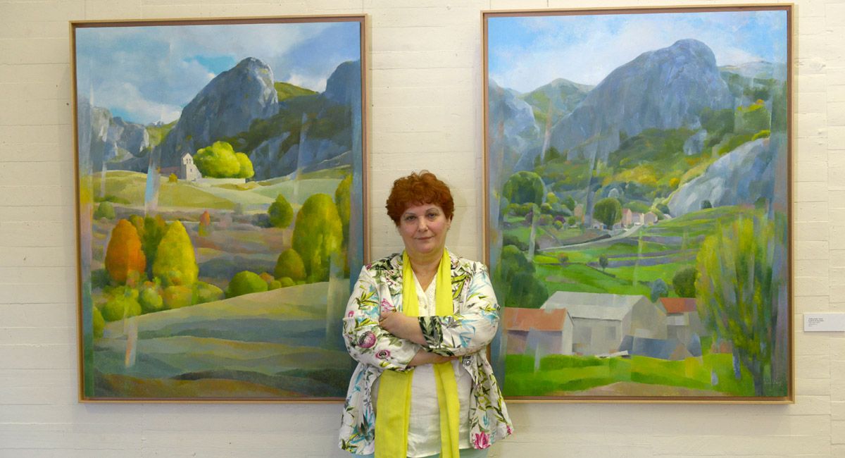 La artista de ascendencia leonesa, Isabel Gutiérrez, posa junto a dos paisajes de su añorado Canseco, en la muestra que acoge el Auditorio. | MAURICIO PEÑA