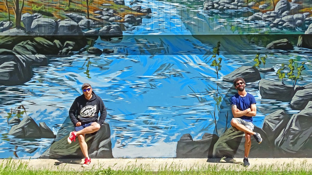 Los artistas Asier Vera y MÜS, junto al mural del río que han pintado en la fachada del pabellón. | Ayto Folgoso